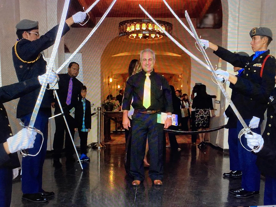 A man wearing black clothing and a gold tie stands beneath raised ceremonial swords at an indoor gathering.
