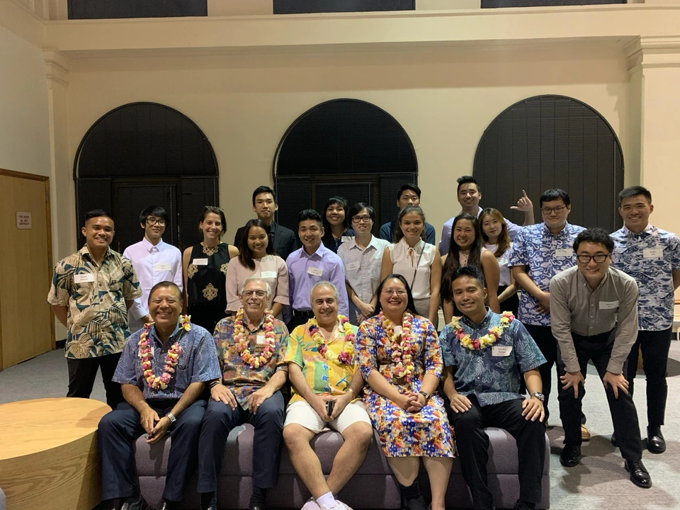 An indoor group photograph with five people seated in the front row wearing flower leis, and others standing behind them.