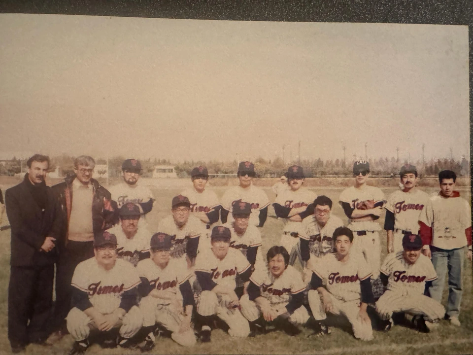 A sports team posing in two rows outdoors in daylight, with several people standing alongside.