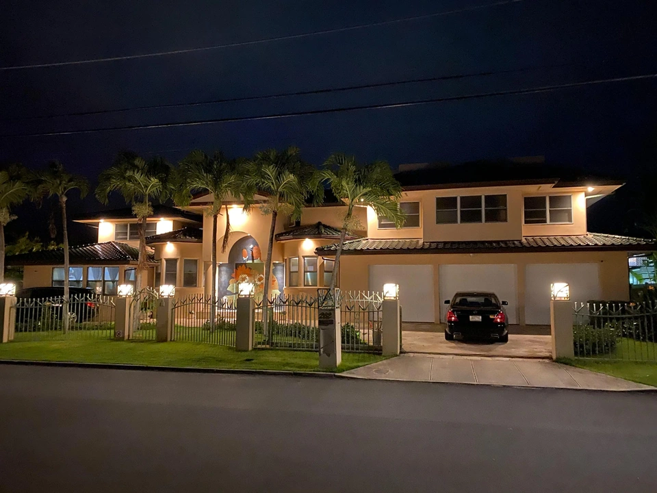 A building exterior at dusk with palm trees, a gated frontage and a parked car.