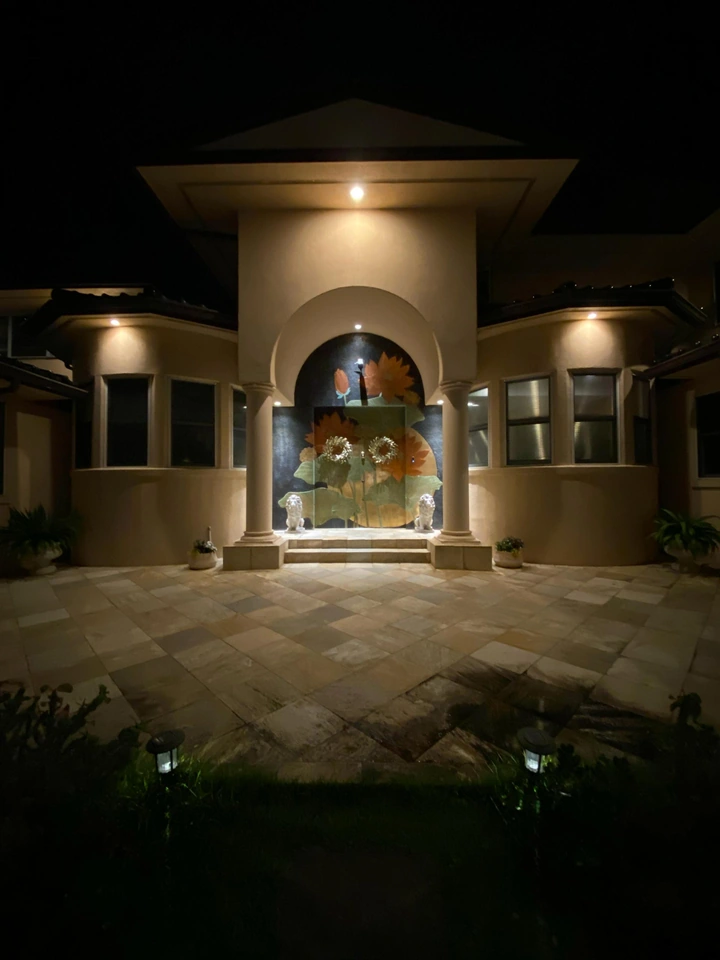 An illuminated entrance at night with columns, a floral door design, steps and a paved courtyard.