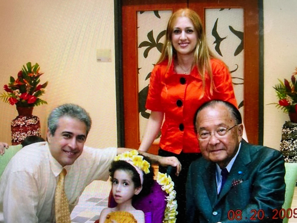 Two seated men, a child and a standing woman pose indoors near floral arrangements.