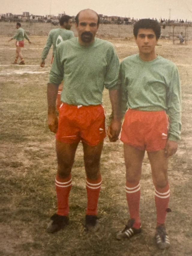 Two people standing on a grass sports field wearing green shirts, red shorts and red socks.
