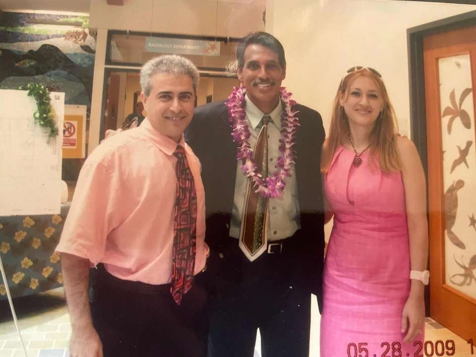 Three adults stand together indoors; the man in the middle wears an orchid lei.