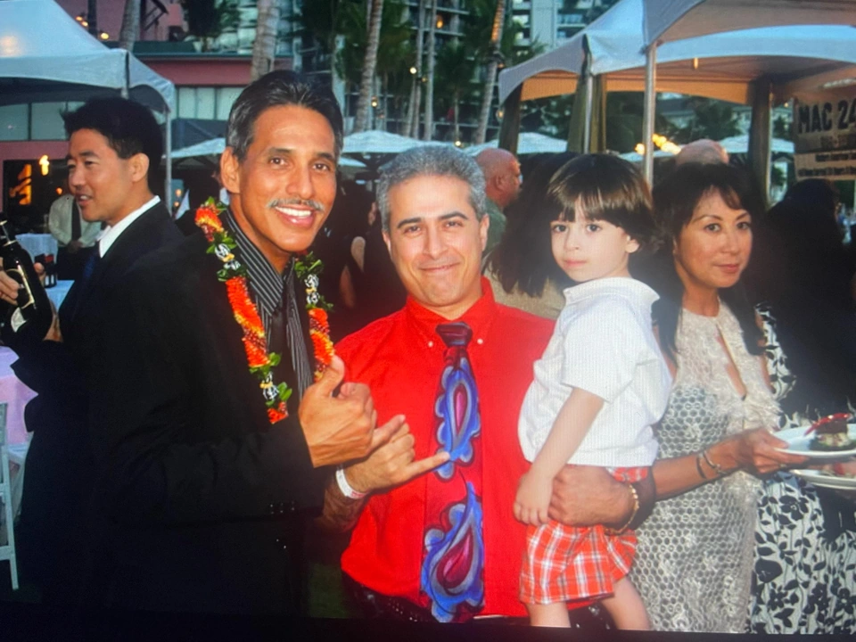 A group poses under outdoor event tents, including a man wearing a flower lei and a man holding a child.
