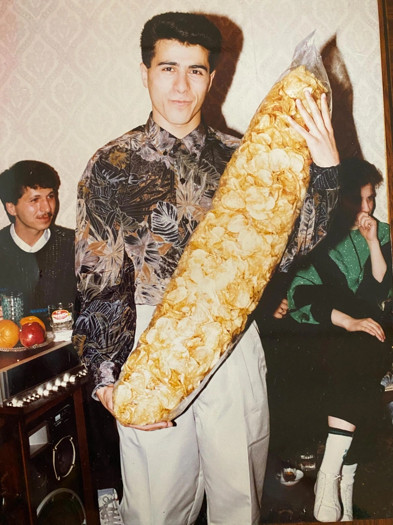 Morad Shaker as a young man holding a large clear bag of potato chips, with two other people seated in the original photograph.