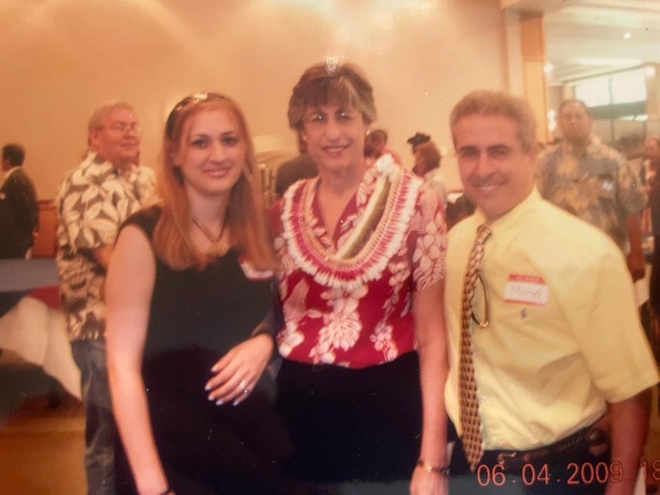 Three guests stand together at an indoor reception; the middle guest wears a flower lei.